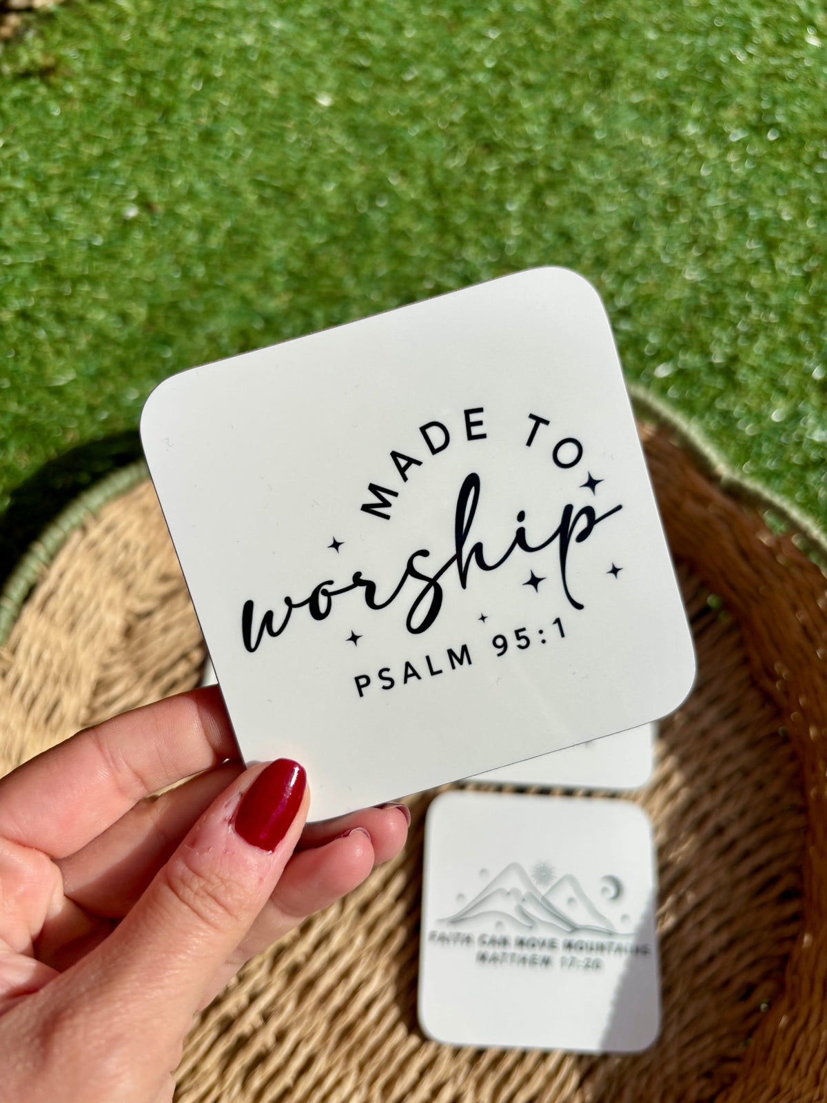 A hand holds a square white coaster featuring black text that reads 'Made to Worship' against a green grassy background and a woven tray.