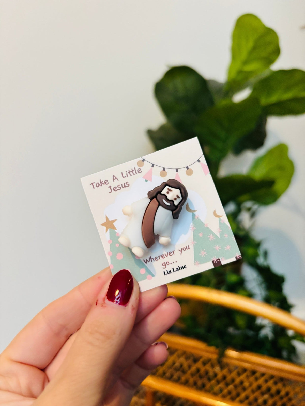 A mini Jeuss figurine on a Christmas design backing card with a plant and wicker basket in background