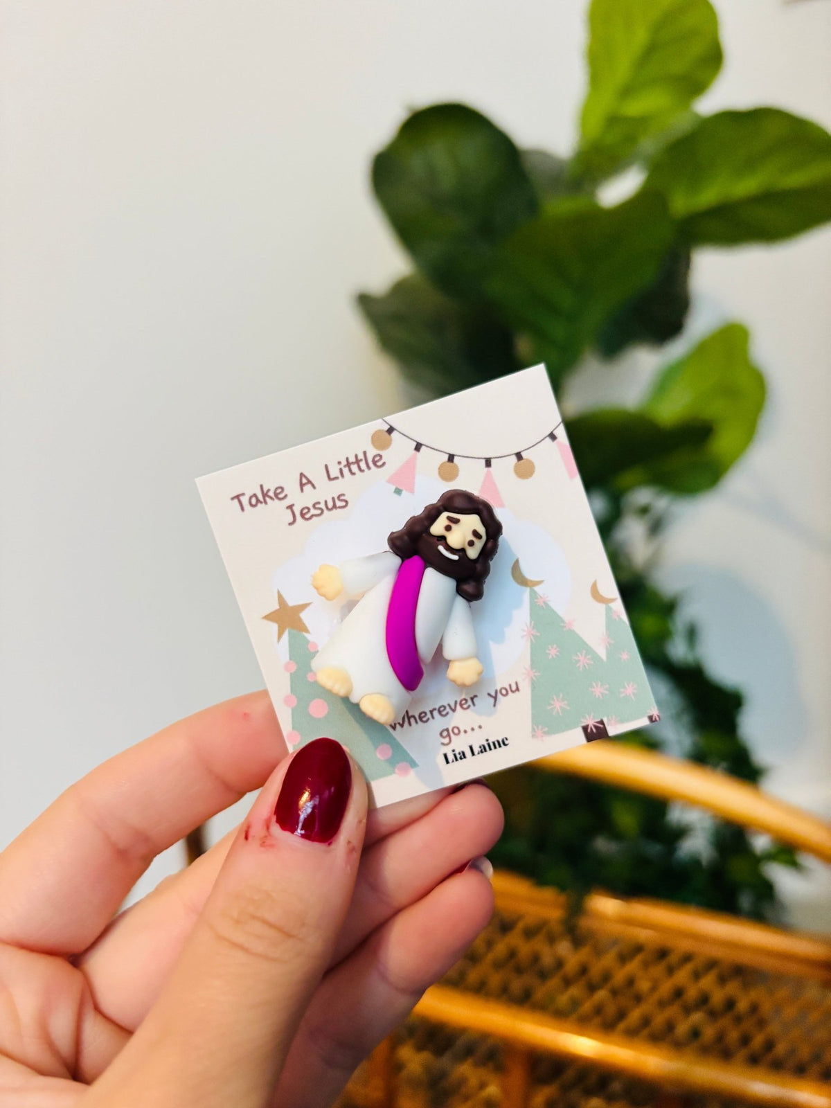 A hand with dark red nail polish holds a small printed card featuring a clay figurine of Jesus with a white robe and purple sash. The card reads 'Take A Little Jesus wherever you go.