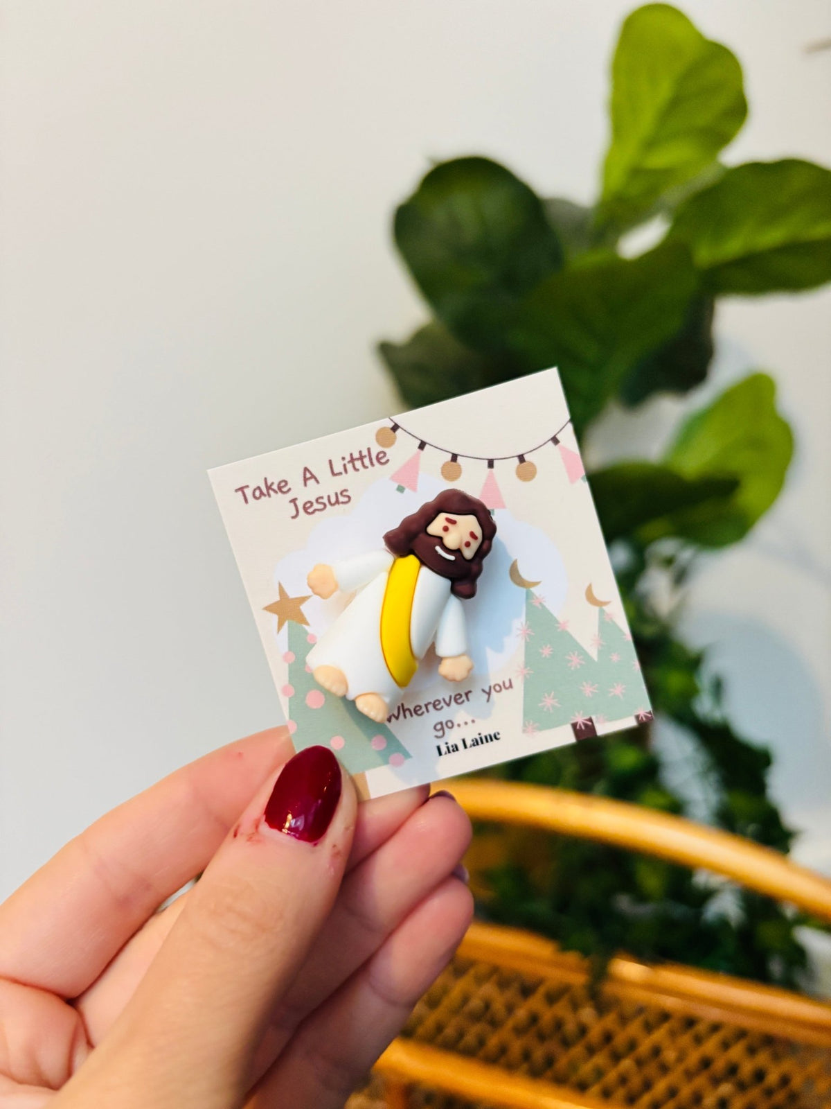 A mini Jeuss figurine on a Christmas design backing card with a plant and wicker basket in background