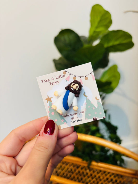 A hand with dark red nail polish holds a small printed card featuring a small Jesus figurine. The card reads 'Take A Little Jesus' and 'Wherever you are, God is near,' with a soft-focus indoor background showing green leaves and a woven chair.