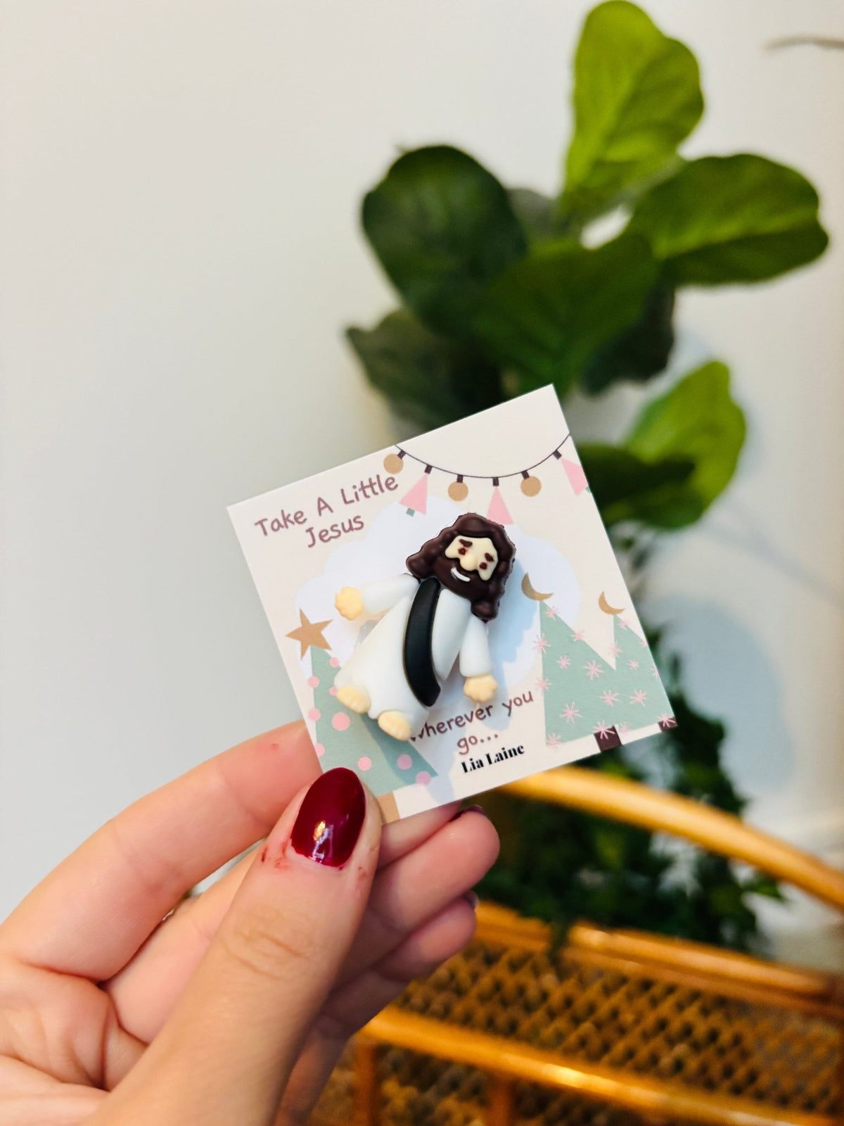 A mini Jeuss figurine on a Christmas design backing card with a plant and wicker basket in background