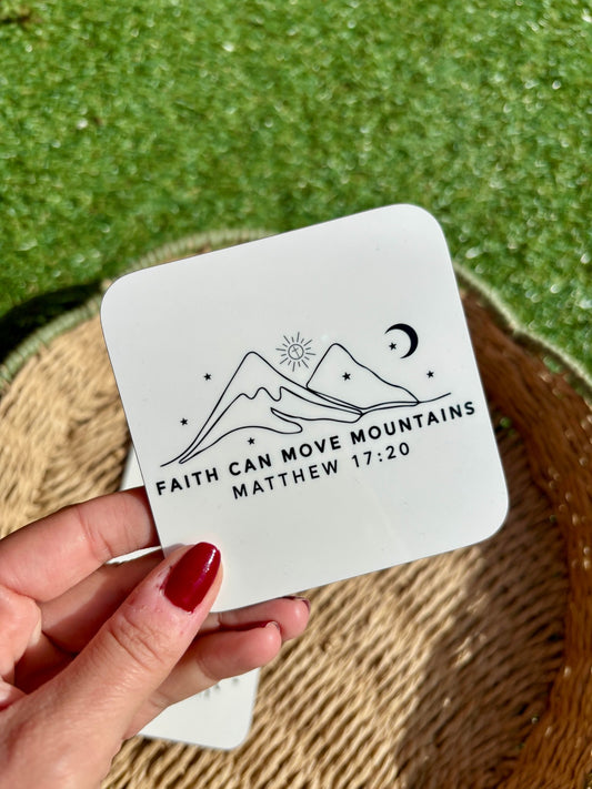 A hand holding a white coaster with black printed text 'Faith Can Move Mountains' on a green grassy background with a woven basket