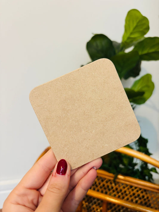 A hand with red nail varnish holding up a coaster showing the back as a brown hardboard, with a plant and wicker basket in the background
