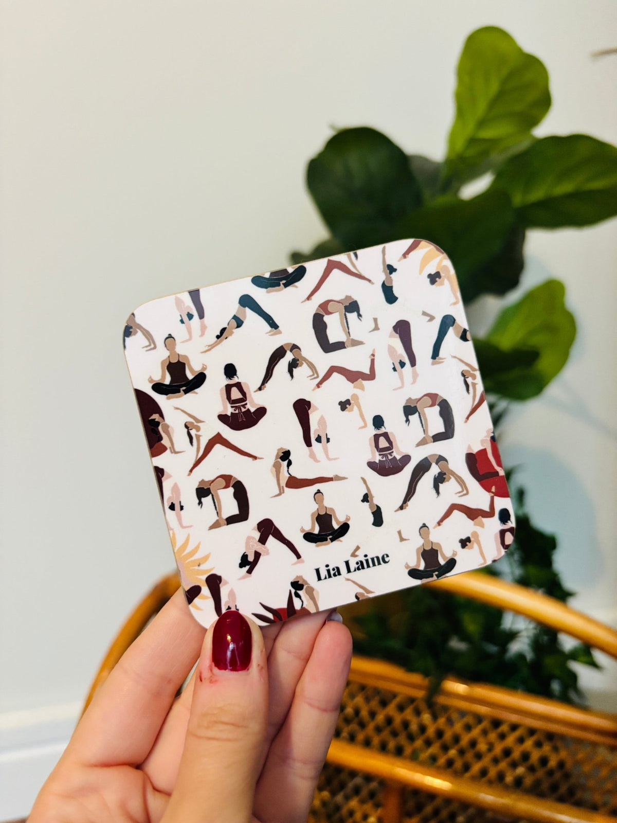 A hand with red nail varnish holding a square coaster with a collage of yoga poses printed on it, with a green plant and wicker basket in the background
