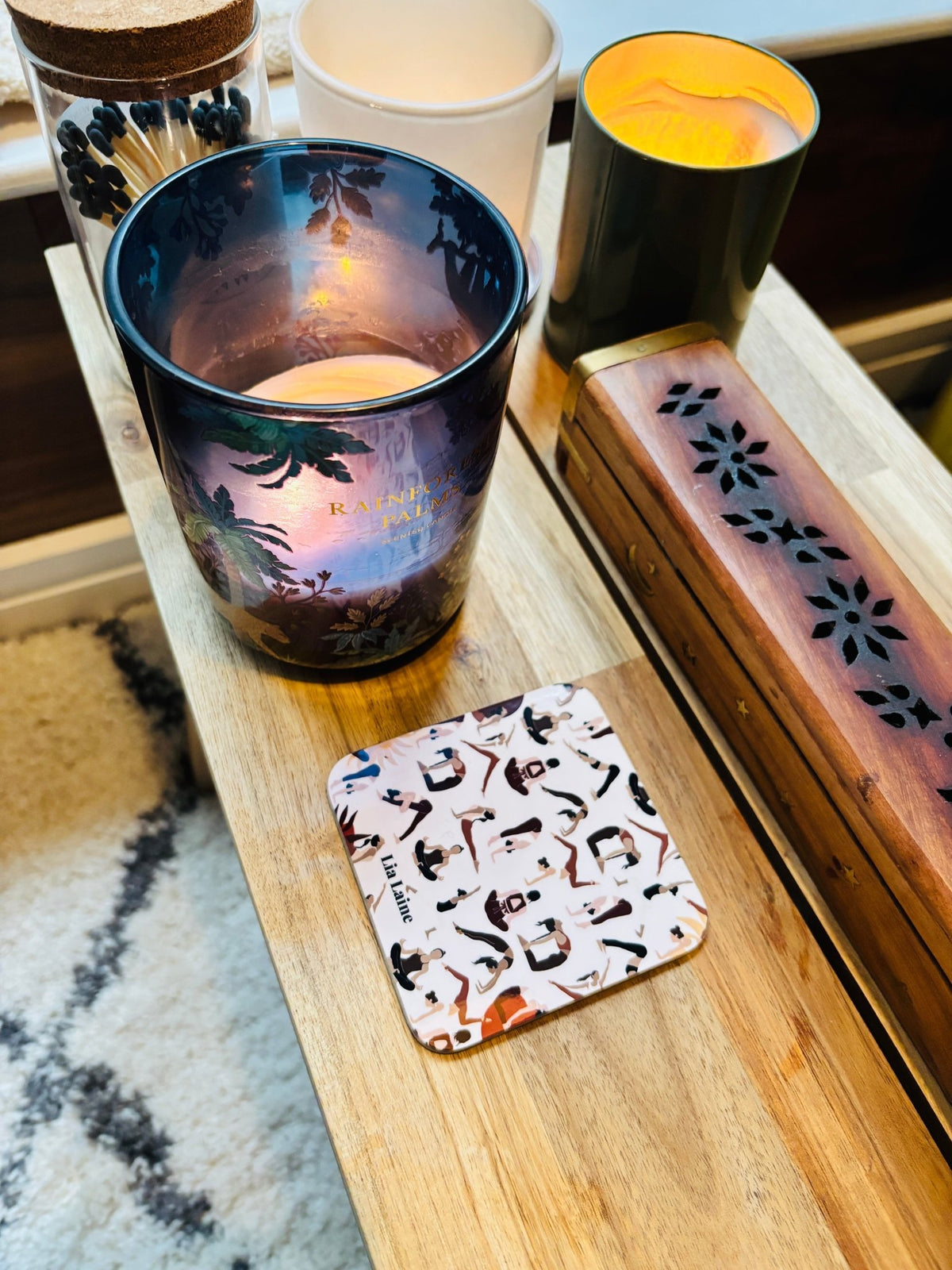 A homely table showing a printed coaster with a collage of yoga poses, next to a collection of candle and insense box on a wooden side table with a cream rug